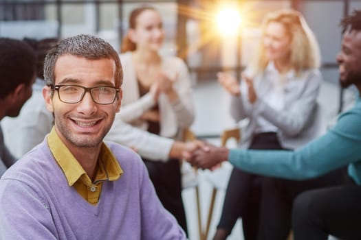 A happy young businessman, in a purple sweater, sits against the background of his colleagues in a modern office