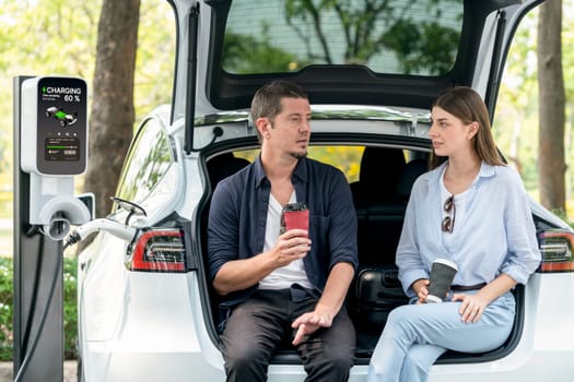 Lovely young couple drinking coffee while recharging battery for electric car during road trip travel EV car in natural forest or national park. Eco friendly travel during vacation and holiday. Exalt