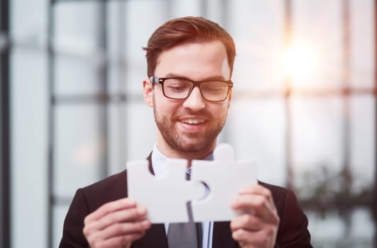 Portrait of an optimistic businessman holding puzzle pieces, solving problems, looking at the camera, wearing a black suit.