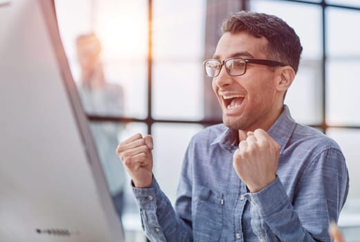 Happy businessman rejoicing success at workplace in office, looking at laptop screen with euphoric expression.