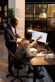 Manager analyzing financial growth report with coworker, working overhours at company strategy planning business investment. African american team discussing marketing statistics in startup office