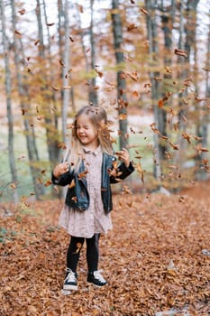 Little laughing girl stands with her eyes closed and her head turned under the falling dry leaves in the autumn forest. High quality photo