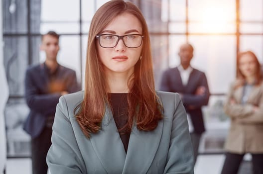 businesswoman standing with her arms crossed in an office while her colleagues have a meeting in the background.