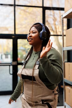 Cheerful manager wearing headphones listening music during work break in storehouse, dancing and having fun. African american employee with industrial overall doing merchandise quality control