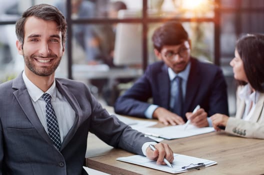 young man posing for the camera while sitting at a table in the office against the background of his colleagues