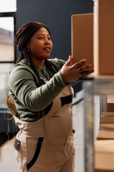 Cheerful manager taking out cardboard box from shelves, preparing customers order during work shift in warehouse. African american employee wearing industrial overall working at products delivery