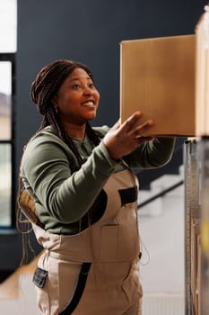 Stockroom worker taking out cardboard box from shelves, preparing customers online orders in warehouse. African american supervisor wearing industrial overall working at products delivery in storehouse
