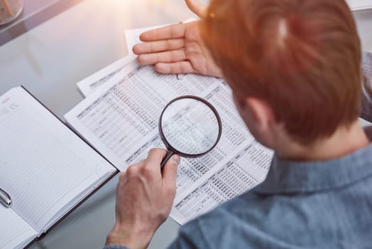 man looks through magnifying glass at document. Concept of examination, verification and problems.