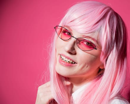 Close-up portrait of a young woman with braces in a pink wig and sunglasses on a pink background