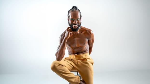 Portrait of smiling African American gay man with bright makeup. Shirtless muscular man sitting on stool and looking at camera. LGBTQ person is posing on white background.