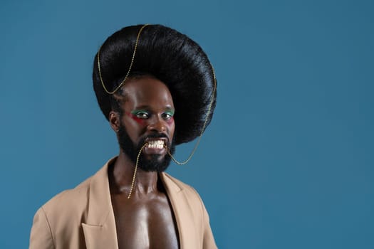 Portrait of African American gay man with makeup and black vintage wig. LGBT person holding gold accessory with teeth and wearing jacket on blue background.