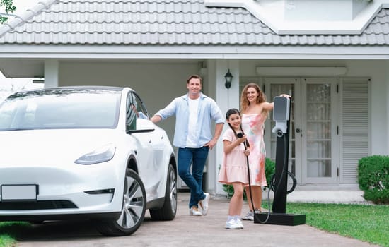 Modern family with environmental conscious recharging EV car with home EV charging station, Happy family with little young girl holding and pointing EV charger at camera. Synchronos