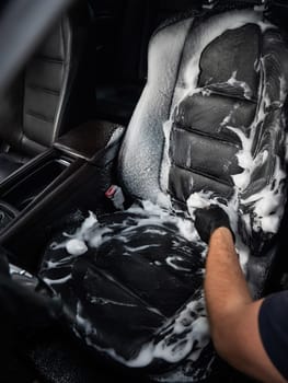 Man cleaning black leather car seat with brush and cleaning foam