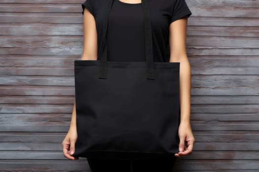 Hand holding black cotton tote bag mockup on a wall background.