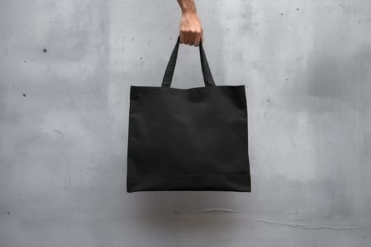 Hand holding black cotton tote bag mockup on a wall background.