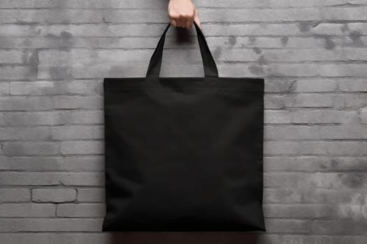 Hand holding black cotton tote bag mockup on a wall background.