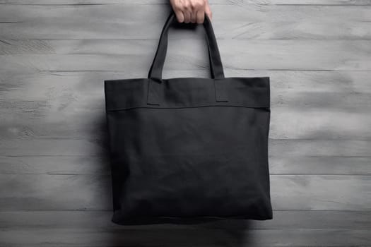 Hand holding black cotton tote bag mockup on a wall background.