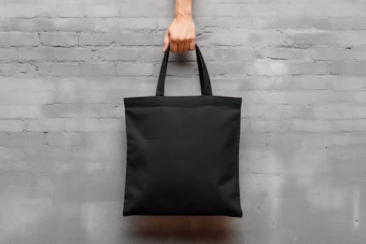 Hand holding black cotton tote bag mockup on a wall background.