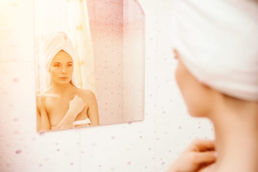 Portrait of young girl with towel on head in white bathroom looks and touches her face in the mirror and enjoys youth and hydration. Natural beauty, home care for problem skin.