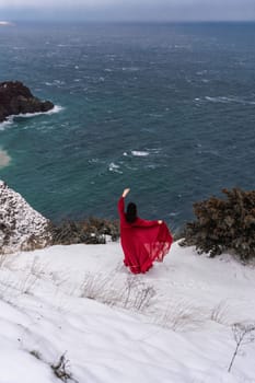 Woman red dress snow sea. Happy woman in a red dress in the snowy mountains by the emerald sea. The wind blows her clothes, posing against sea and snow background