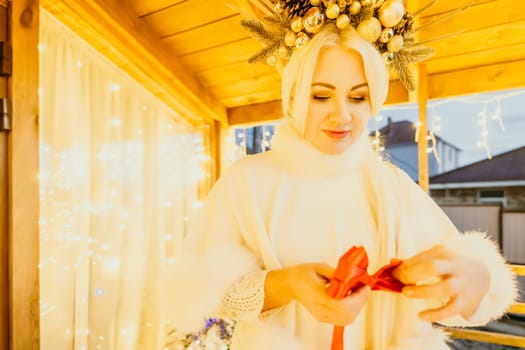 A woman in a white dress is holding a gold box with a red ribbon. She is wearing a crown on her head. The scene takes place in a room with a door and a window. The woman appears to be opening the gift box.