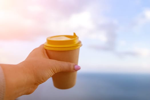 Hand holding Yellow cup with lid, coffee against a backdrop of a blue sky and sea. Illustrating cup and beverage.