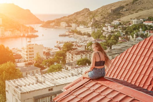 Woman sits on rooftop, enjoys town view and sea mountains. Peaceful rooftop relaxation. Below her, there is a town with several boats visible in the water. Rooftop vantage point