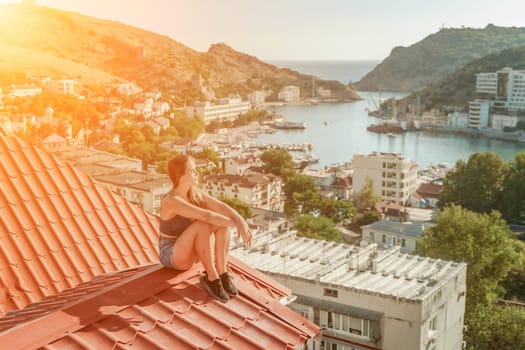 Woman sits on rooftop, enjoys town view and sea mountains. Peaceful rooftop relaxation. Below her, there is a town with several boats visible in the water. Rooftop vantage point