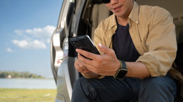 Young solo traveler sitting on the open trunk and using mobile phone over blue sky. Travel lifestyle concept.