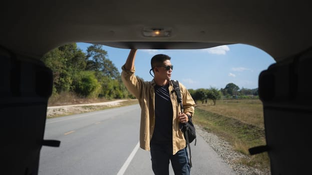 Solo travel man carrying backpack standing on countryside road a seen through open vehicle trunk.