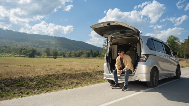 Young male solo traveler sitting on the open trunk of car with beautiful mountain views and blue sky.