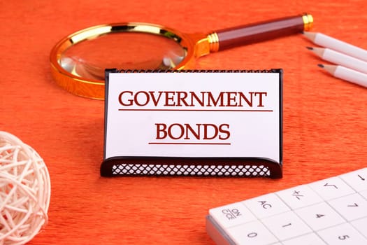 Government Bonds text on a white business card on a stand on an orange background, next to a magnifying glass, a calculator and pencils