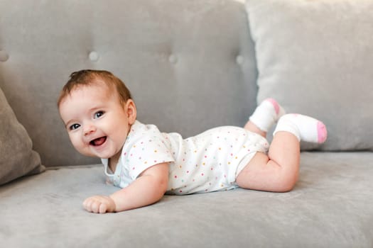 Cheerful cute toddler in short sleeve bodysuit and socks lying on couch and laughing while looking at camera at home