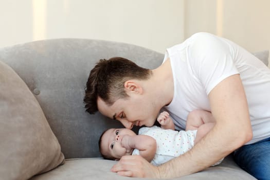 Side view of happy caring father in casual clothes looking down while sitting on couch with pillows and touching baby cheek with nose in living room at home