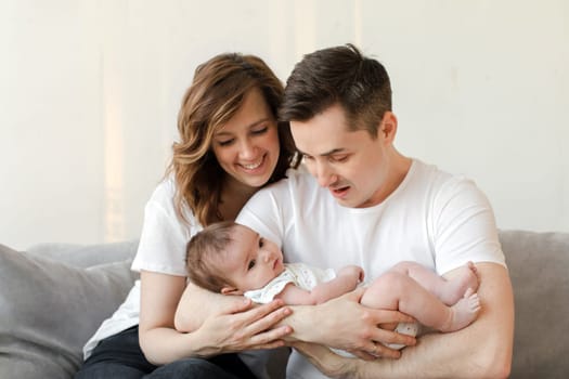 Positive parents in casual outfit sitting on gray couch and cuddling adorable little child while spending time together at home during weekend against beige wall on background