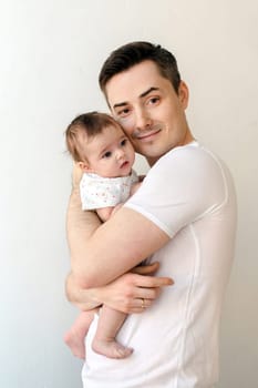 Cheerful man in white t shirt smiling and embracing adorable little child while standing in apartment and looking at camera against beige wall on background