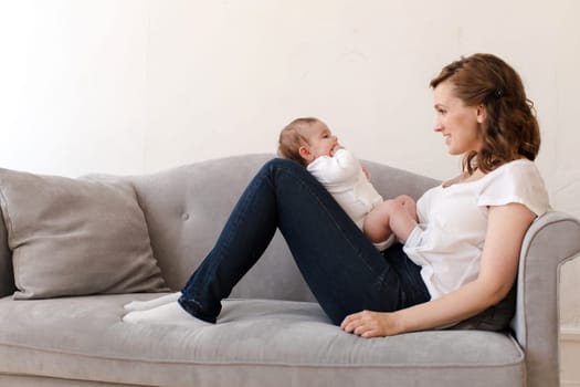 Side view of smiling young mother in casual clothes looking away while relaxing on sofa and playing with child on folded legs in cozy living room at home