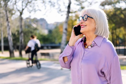senior woman smiling happy talking by mobile phone while walking at park, concept of technology and elderly people leisure