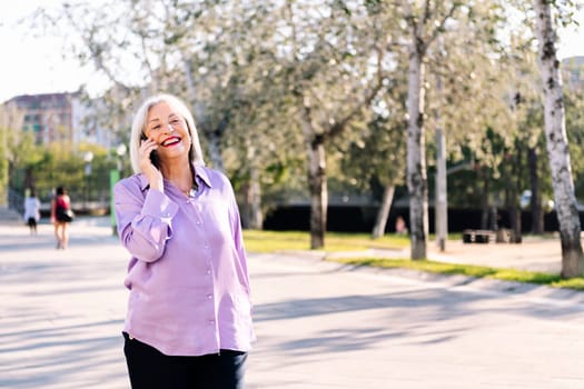 senior woman laughing happy talking by mobile phone while walking at park, concept of technology and elderly people leisure, copy space for text