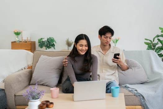 Happy young couple using mobile phone and laptop relax on sofa in living room watch video on laptop together.
