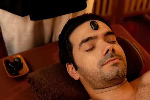 Caucasian man enjoying relaxing anti-stress head massage with hot stone and pampering facial beauty skin recreation leisure in warm candle light ambient salon spa in resort or hotel. Quiescent