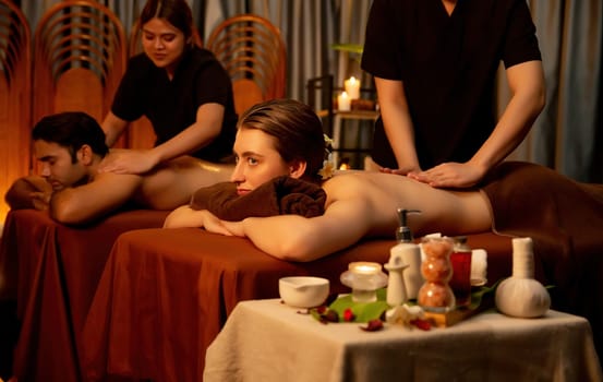 Caucasian couple customer enjoying relaxing anti-stress spa massage and pampering with beauty skin recreation leisure in warm candle lighting ambient salon spa at luxury resort or hotel. Quiescent