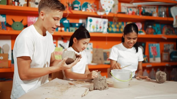 Smart highschool student using hand to press on clay piece at pottery workshop. Group of happy diverse children working or modeling cup of clay with dough and equipment scatter around. Edification.