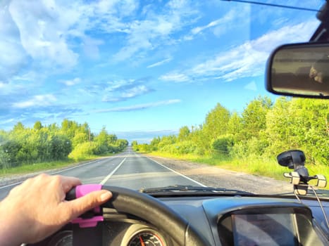 Car salon, windshield, hand of woman on steering wheel and landscape. View from seat of driver on nature with Road, trees, blue sky at a sunny day. Single trip of female traveler. Partial focus