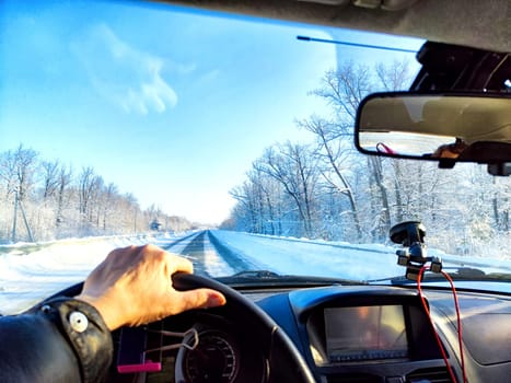 Hand of woman on the steering wheel in car and view through the windshield of a winter road with snow-covered trees