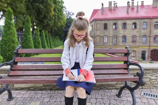 Back to school. Outdoor portrait of beautiful blond girl 9, 10 years old with backpack writing in notebook. Little student sitting on bench near school building