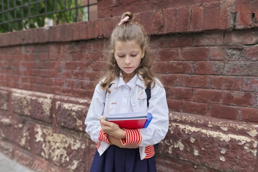 Back to school. Outdoor portrait of beautiful blond girl 9, 10 years old with backpack, notebooks near brick wall of fence school building