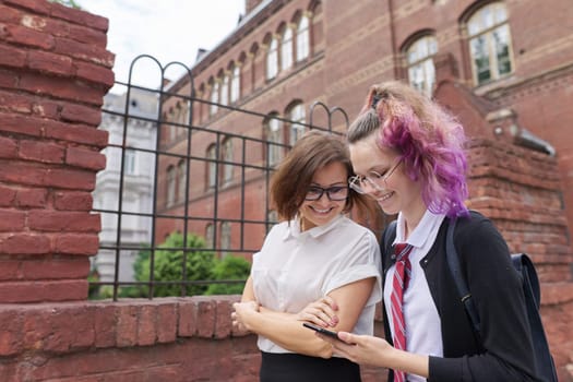 Female student teenager in school uniform with backpack walking and talking with woman teacher, girl showing on smartphone screen, school building background