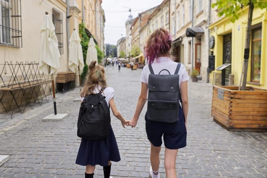 Children going to school, two girls sisters teenager and elementary school student with backpacks walking together, holding hands, back view