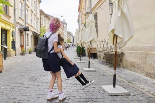 Two girls sisters having fun on the way to school. Teenager girl is circling younger, children are laughing, sunny summer day in city